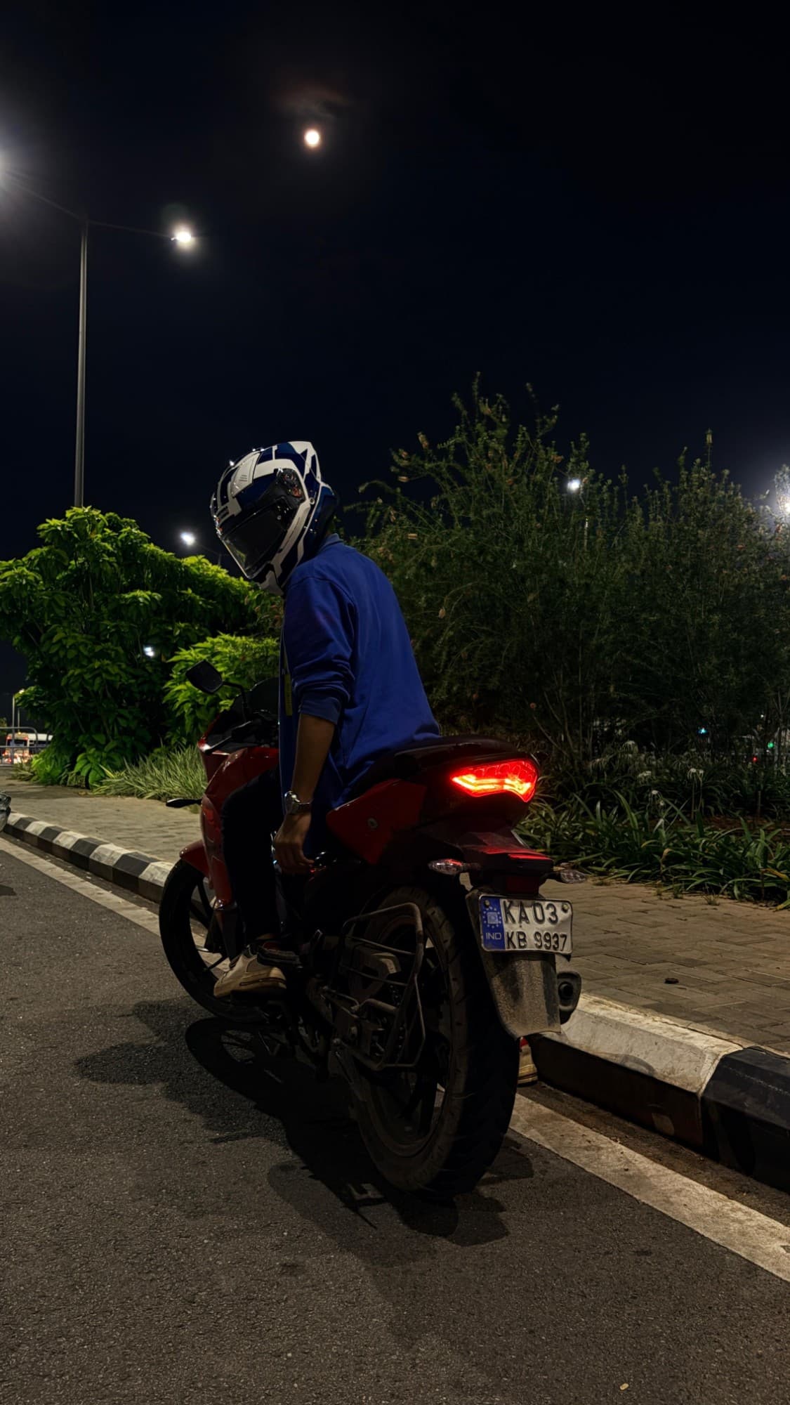 A red motorcycle parked at night on a Bengaluru street, its tail light glowing under a streetlamp and a full moon.