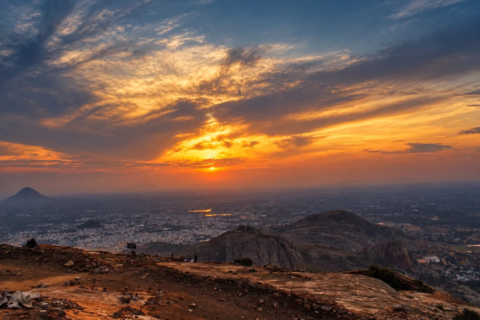Wide sunset over a rocky landscape overlooking the city, with layered mountains and a bright horizon.
